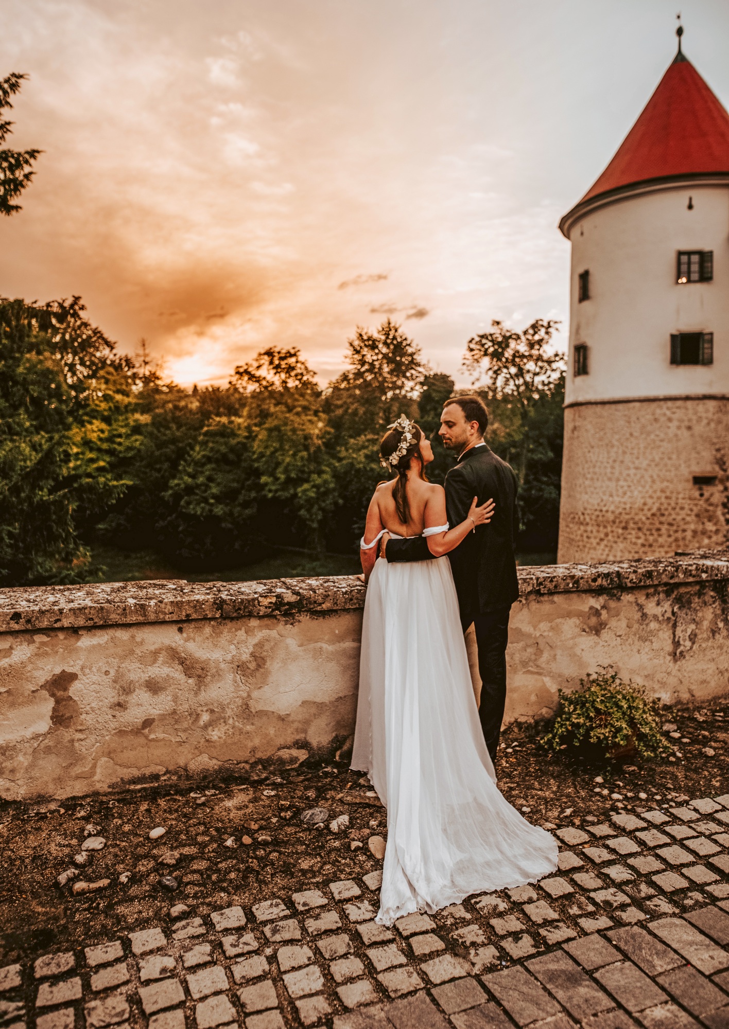 Rear view of bride and groom their wedding day, standing on castle bridge in sunset light.