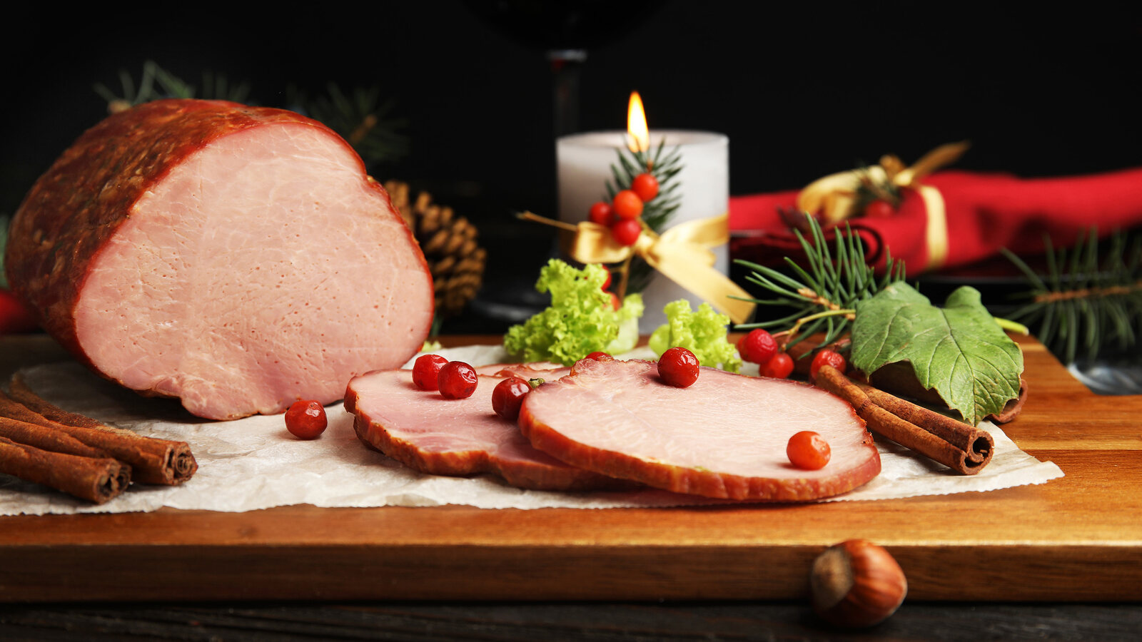 Skivad julskinka på skärbräda med röda bär, kanelstänger och ett tänt ljus
